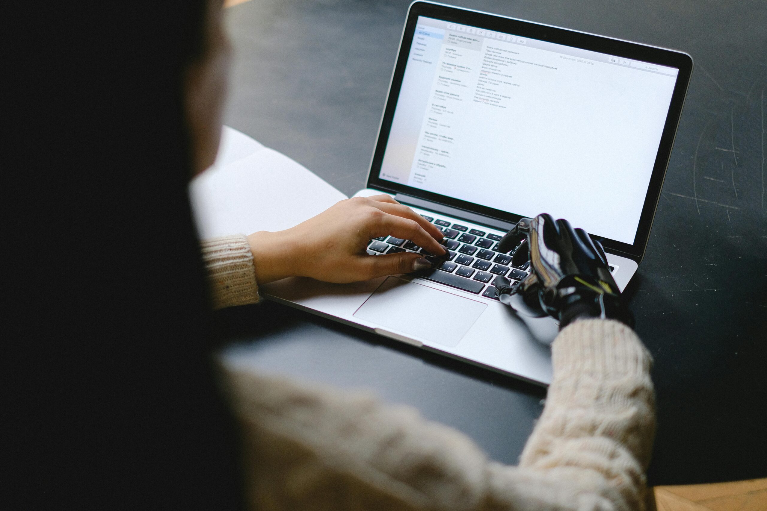 A person wearing a beige sweater types on a laptop, using one natural hand and one prosthetic hand. The laptop screen displays an email inbox, and the scene is set at a dark table in a softly lit environment.<br />
Image by Anna Shvets